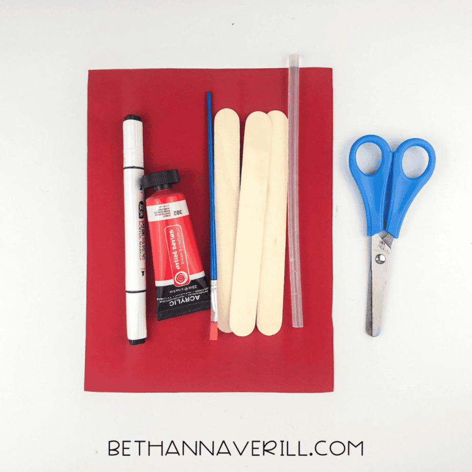 Craft supplies including red paper, popsicle sticks, scissors, paint, brush, marker, and glue stick on a white background