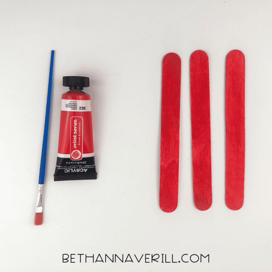 Paintbrush and red paint next to three painted red popsicle sticks