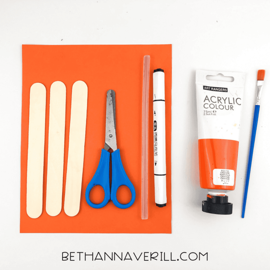 Popsicle sticks, scissors, glue stick, marker, straw, paint, and paper arranged on an orange sheet