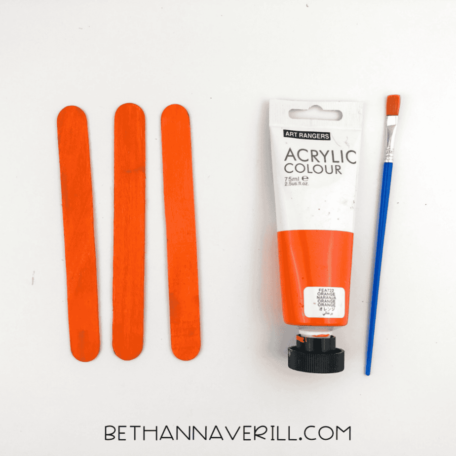 Three popsicle sticks painted orange next to a tube of acrylic paint and a paintbrush