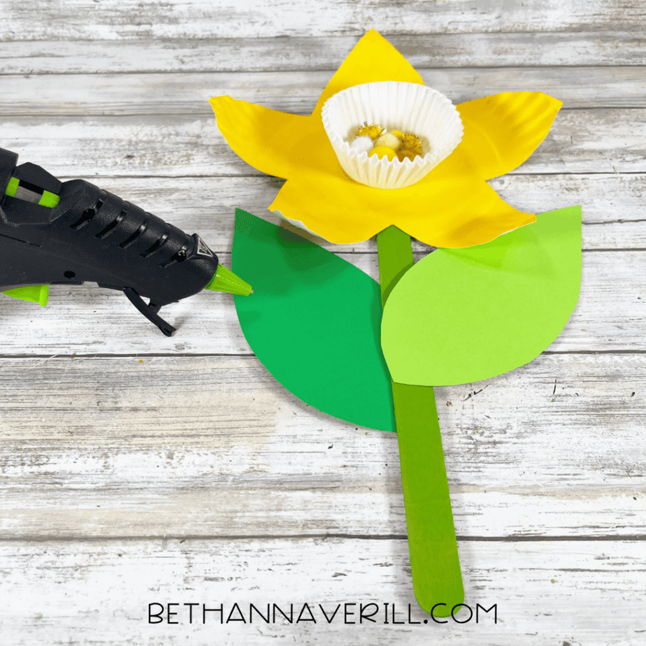 Paper leaves being glued to a green craft stick stem with a yellow flower attached.