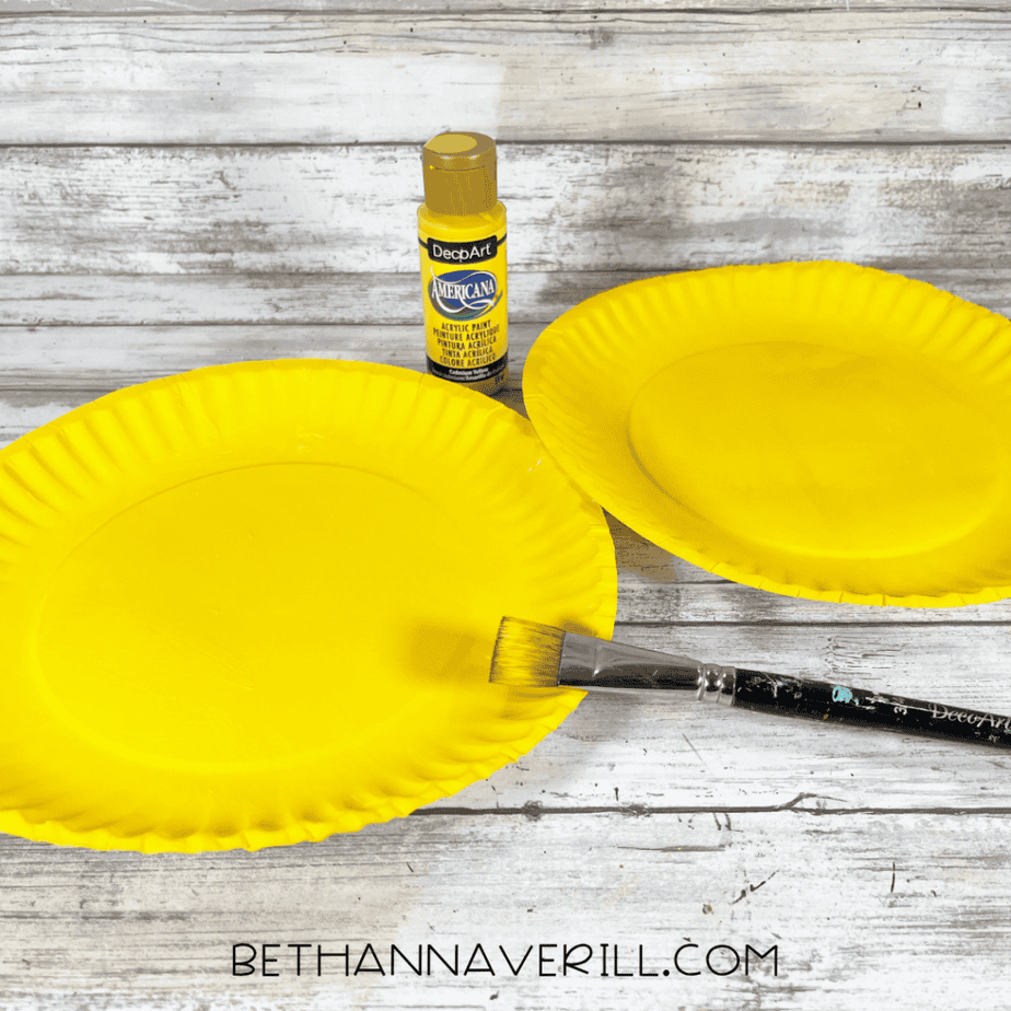 Two paper plates painted bright yellow with a paintbrush and yellow acrylic paint nearby.