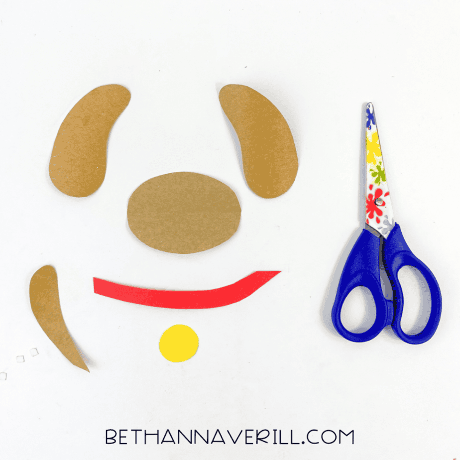 Cut paper pieces for a popsicle stick dog craft including ears, muzzle, tail, collar strip, and circle cut from colored cardstock with scissors nearby.