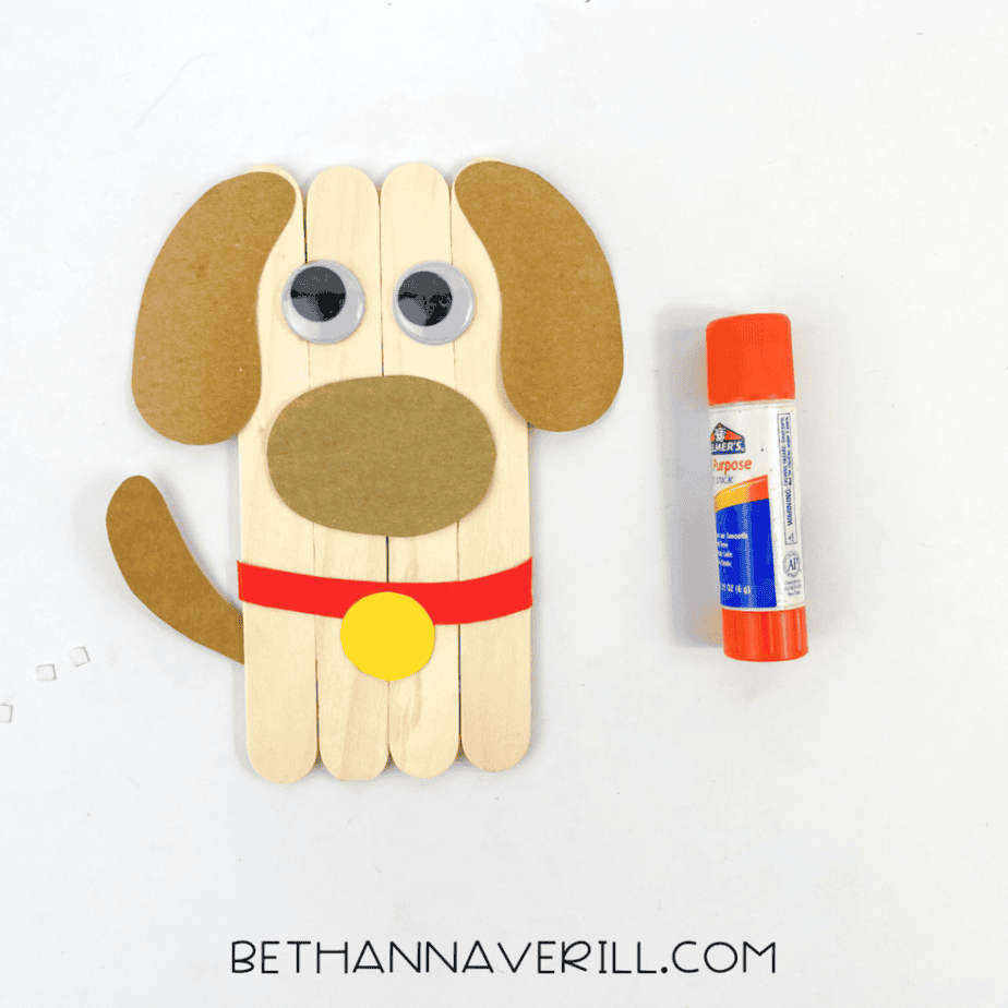 Partially assembled dog popsicle stick craft with googly eyes, paper ears, muzzle, collar, and tail attached to wooden craft sticks.