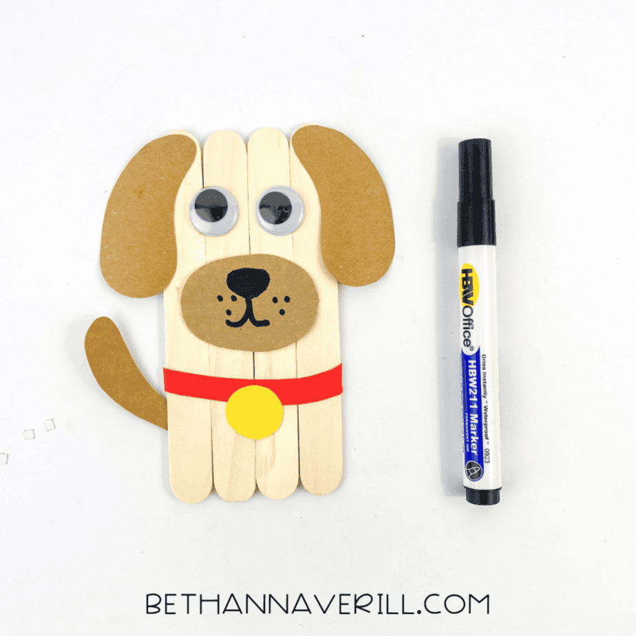 Dog popsicle stick craft with facial details drawn on the muzzle using a black marker, shown beside the marker.