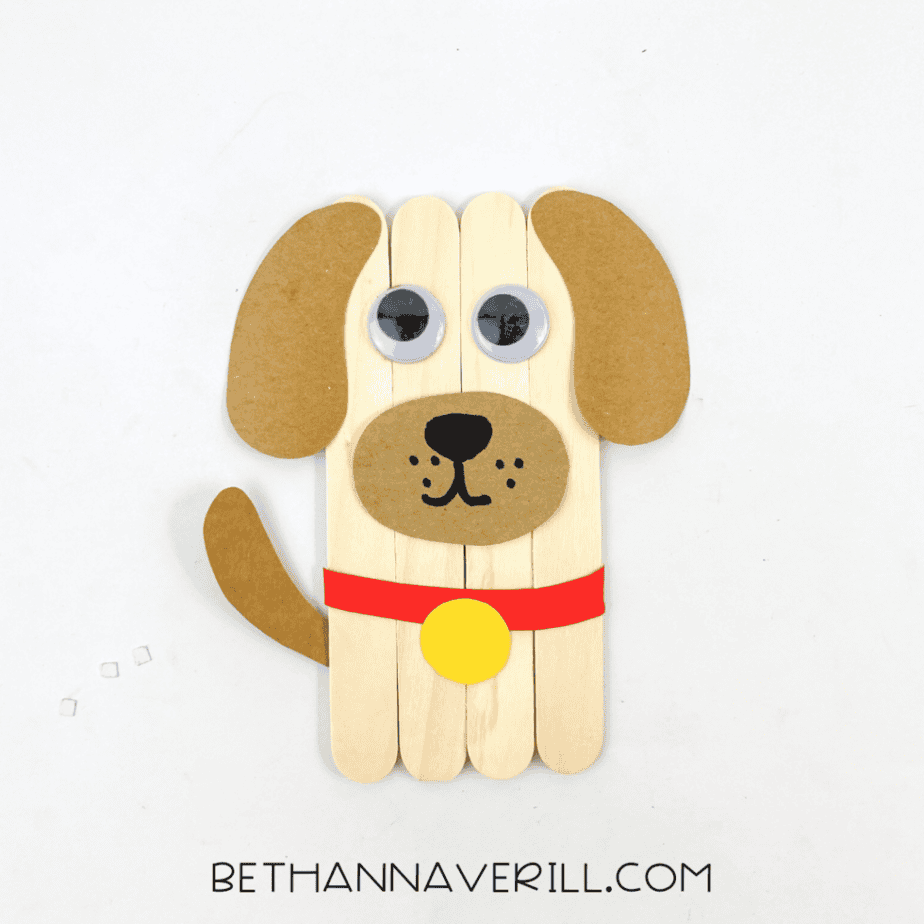 Finished dog popsicle stick craft made with wooden craft sticks, paper ears, googly eyes, and a red paper collar with a yellow circle tag.