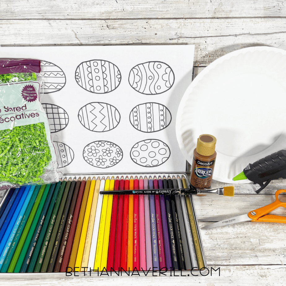 Craft supplies including colored pencils, paper plate, paint, scissors, glue gun, decorative shred, and printed Easter egg coloring page on a wood surface