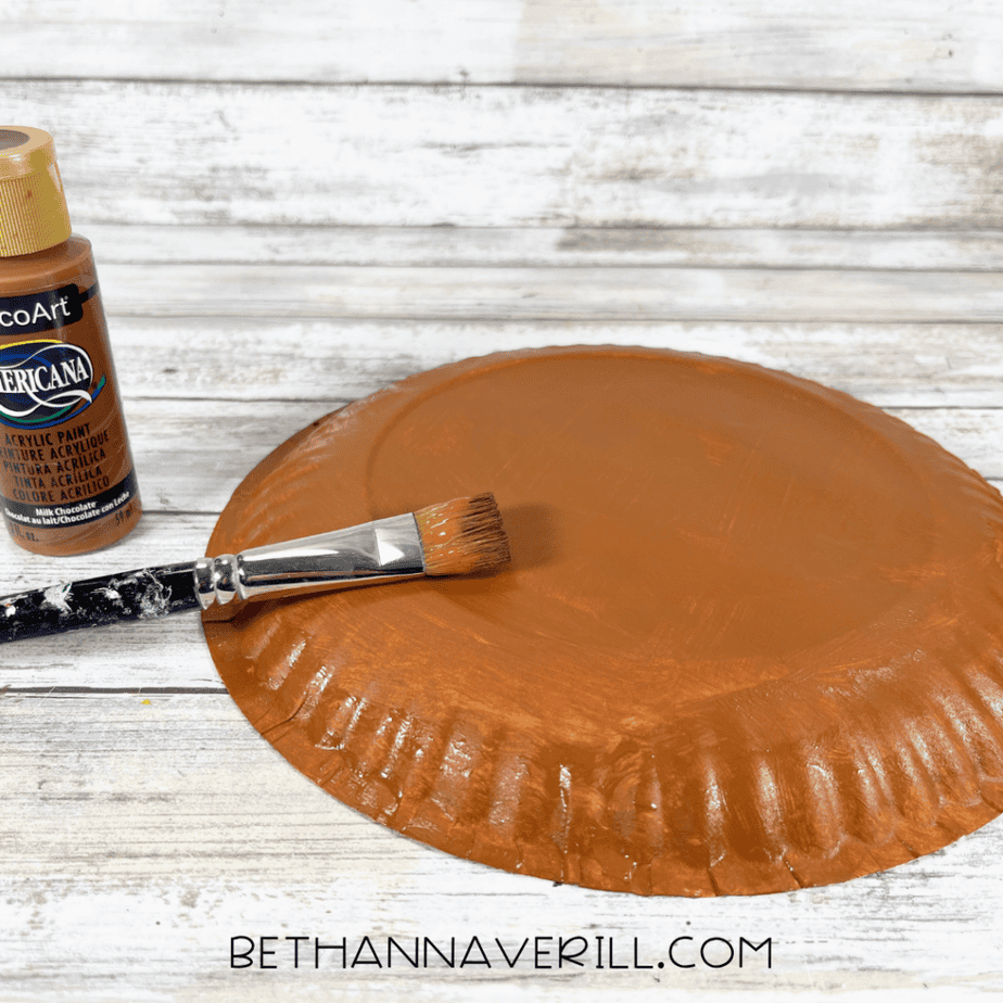Paper plate painted brown with a paintbrush and acrylic paint bottle on a wood background