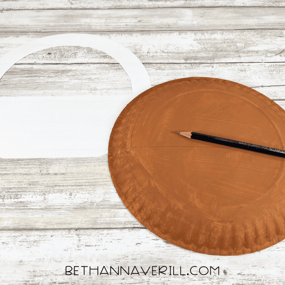 Brown painted paper plate with a pencil drawing a curved outline next to a cut paper template