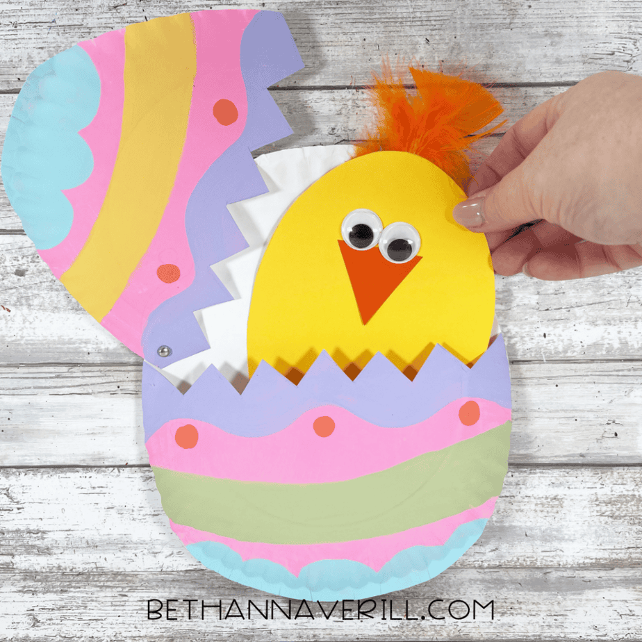 A yellow chick with googly eyes and an orange feather tuft being placed inside the egg shell.