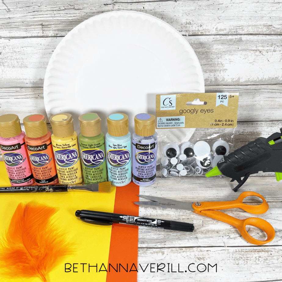Craft supplies arranged on a table: paper plate, acrylic paint bottles, paintbrush, scissors, glue gun, googly eyes, colored paper, marker, and an orange feather.