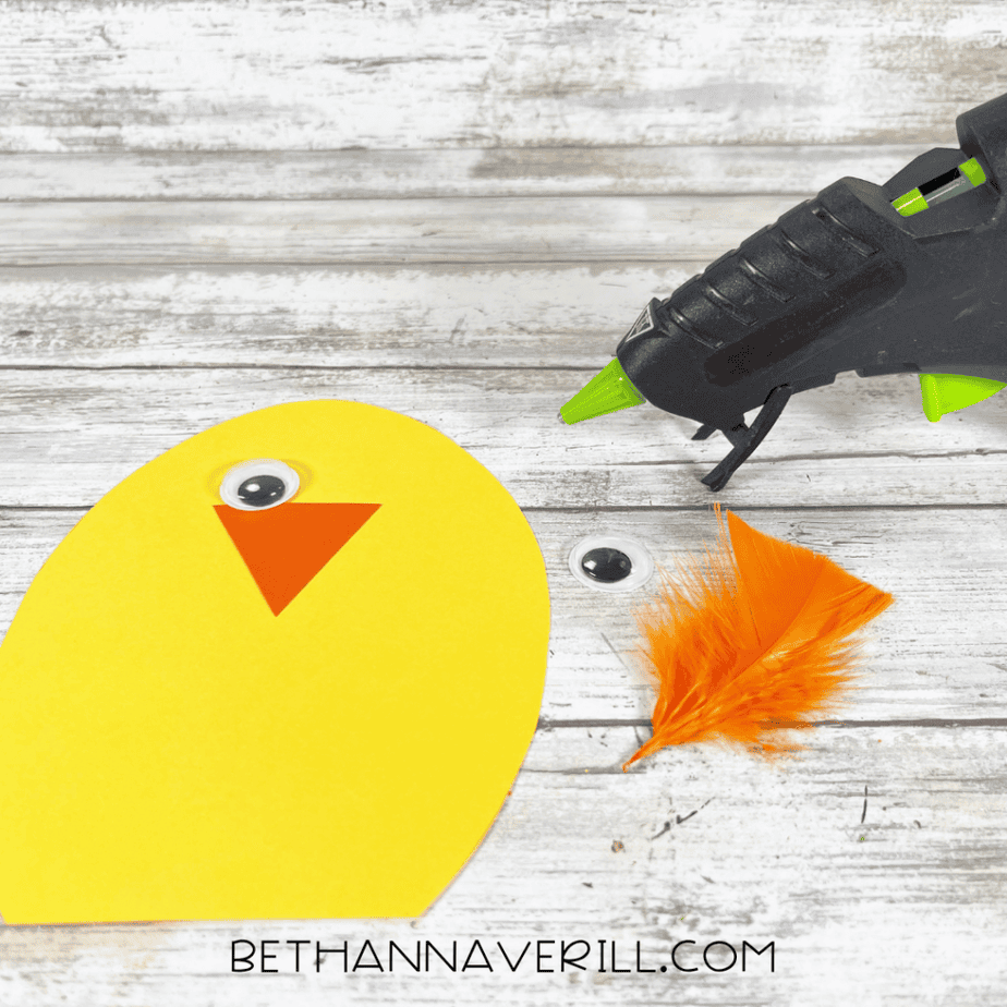 A yellow paper egg shape with a single googly eye and an orange triangle beak being assembled next to a glue gun and feather.