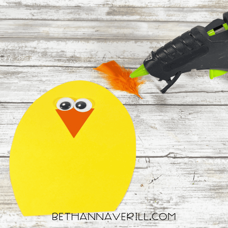 The same yellow egg now with two googly eyes; a glue gun is positioned near an orange feather for attachment.