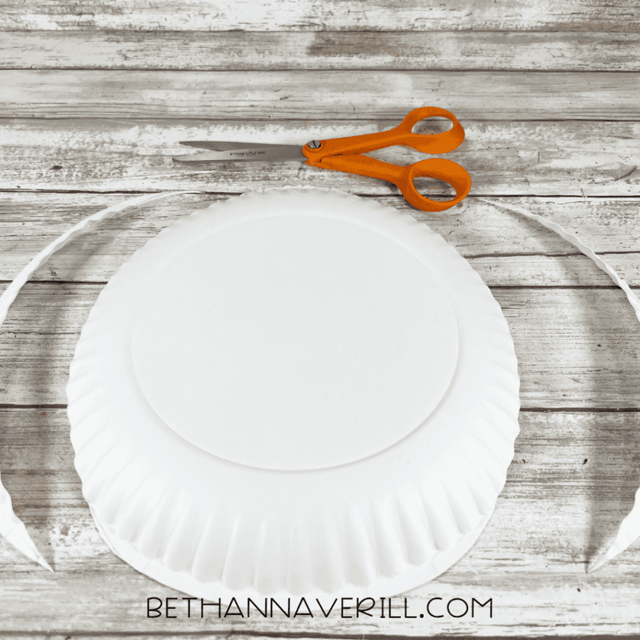 A paper plate with its outer rim cut away, leaving a smooth circular center; scissors are above it.