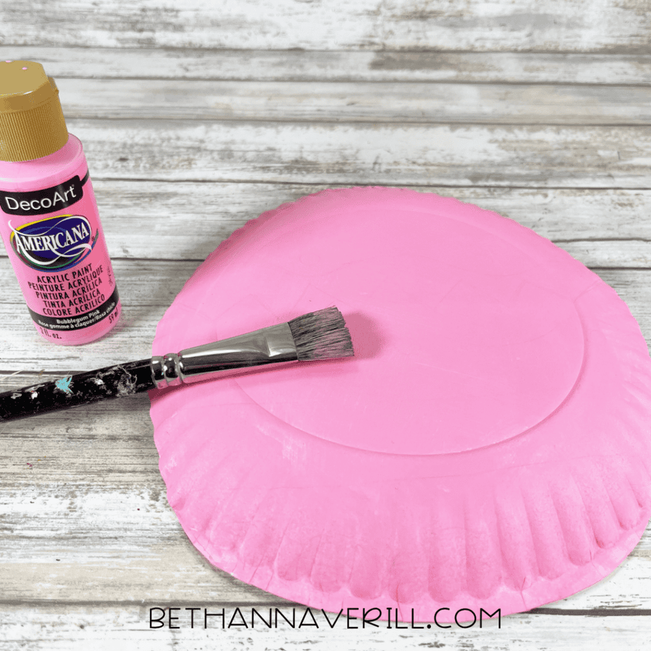 The back of a paper plate being painted pink with a paintbrush and a bottle of pink acrylic paint nearby.