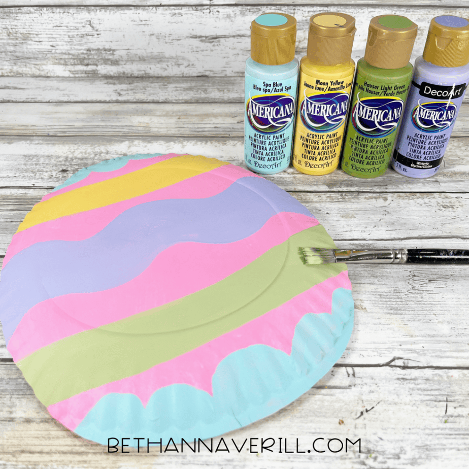 A painted paper plate decorated with horizontal pastel stripes in blue, yellow, pink, purple, and green.