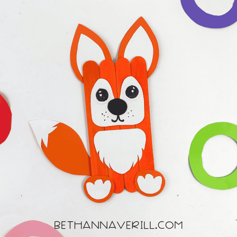 Finished fox popsicle stick craft displayed on a white background surrounded by colorful craft paper shapes.