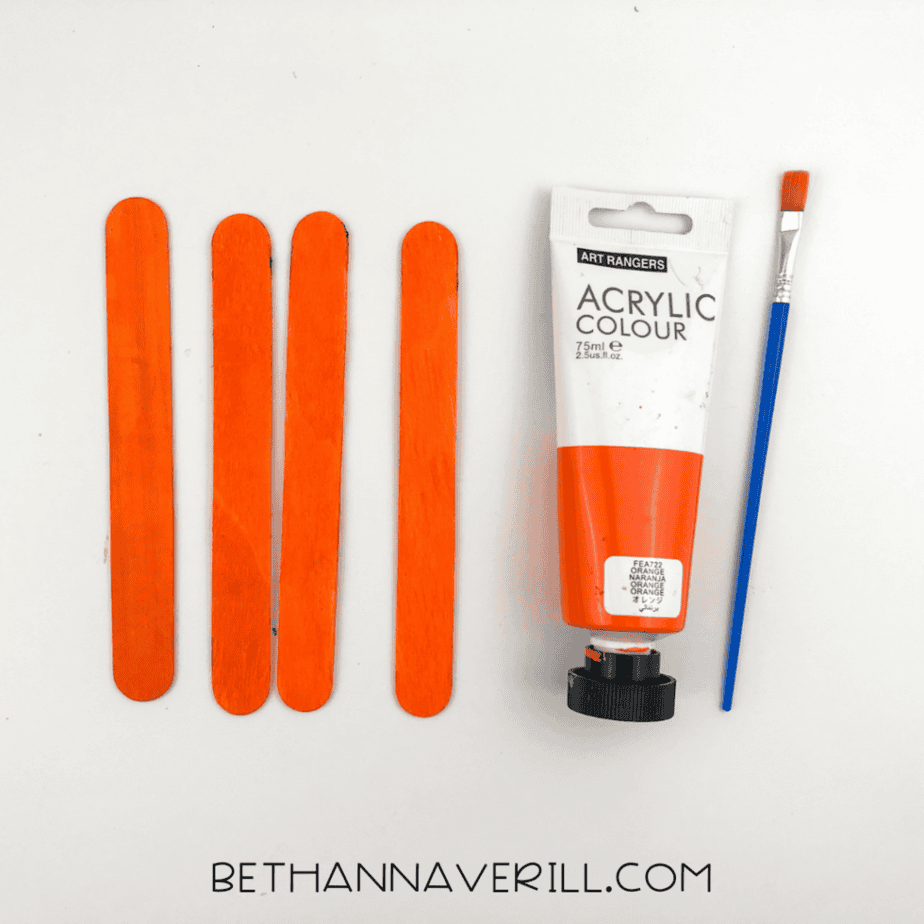 Wooden craft sticks painted orange next to orange acrylic paint and a paintbrush for a fox popsicle stick craft.