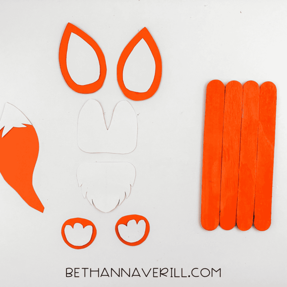 Cut paper pieces arranged beside painted craft sticks showing parts for a fox popsicle stick craft including ears, paws, and tail.