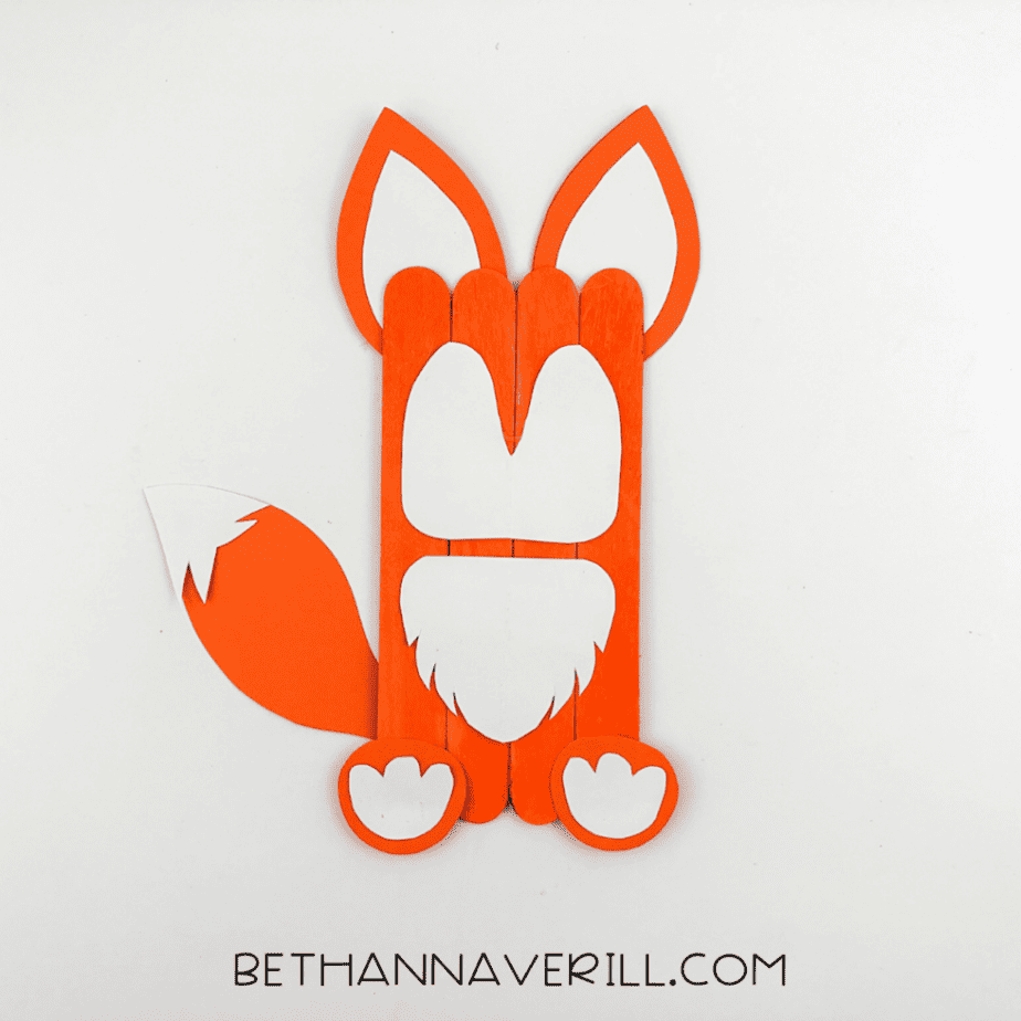 Partially assembled fox popsicle stick craft with orange body, white chest, tail, and paper ears attached.