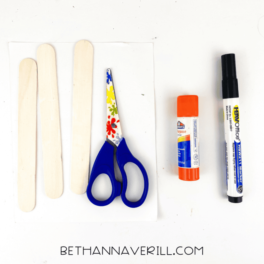 Craft supplies for a ghost popsicle stick craft including wooden craft sticks, scissors, glue stick, black marker, and white cardstock.