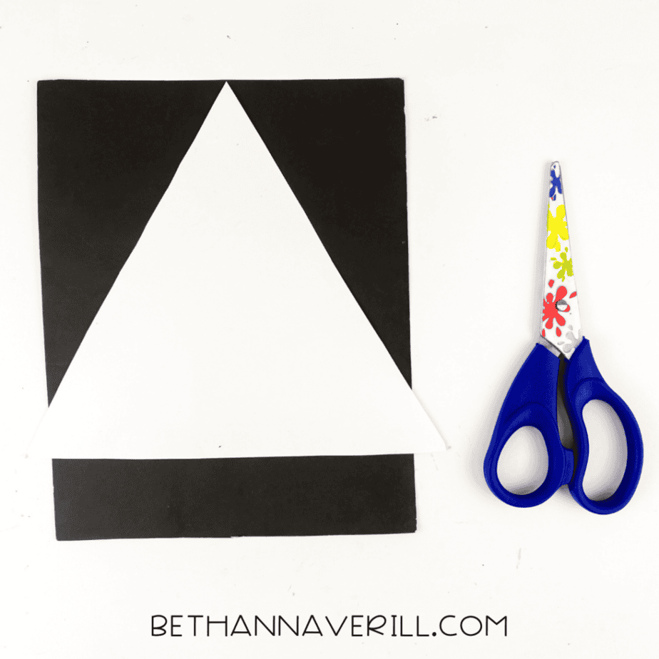 White triangle paper template placed on black cardstock with scissors to cut shapes for a ghost popsicle stick craft.