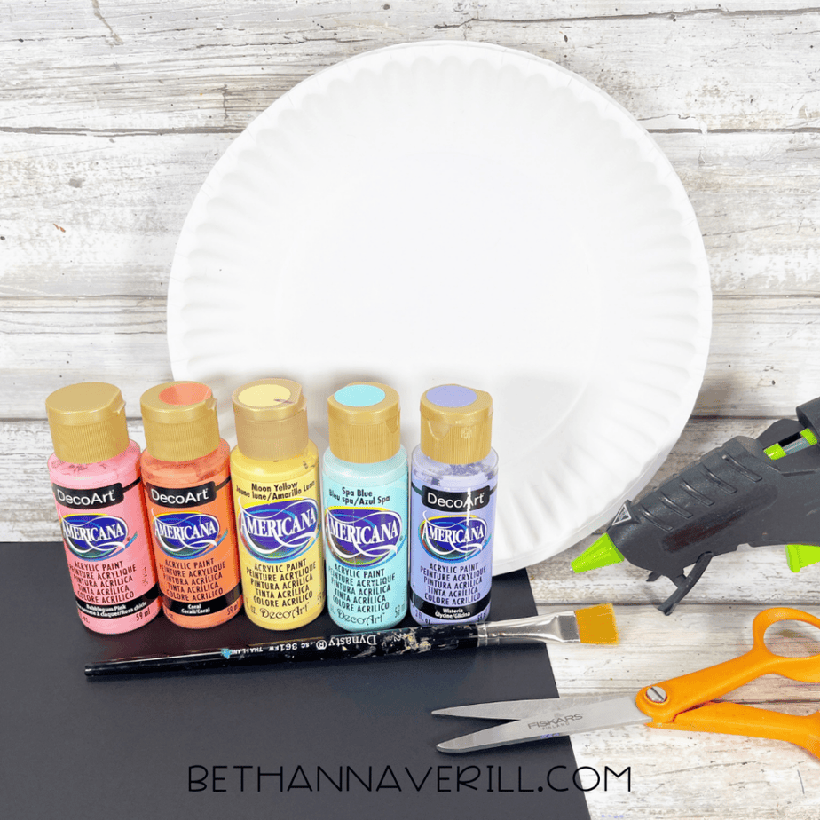 White paper plate with acrylic paint bottles, brush, scissors, glue gun, and black paper on a wood surface.