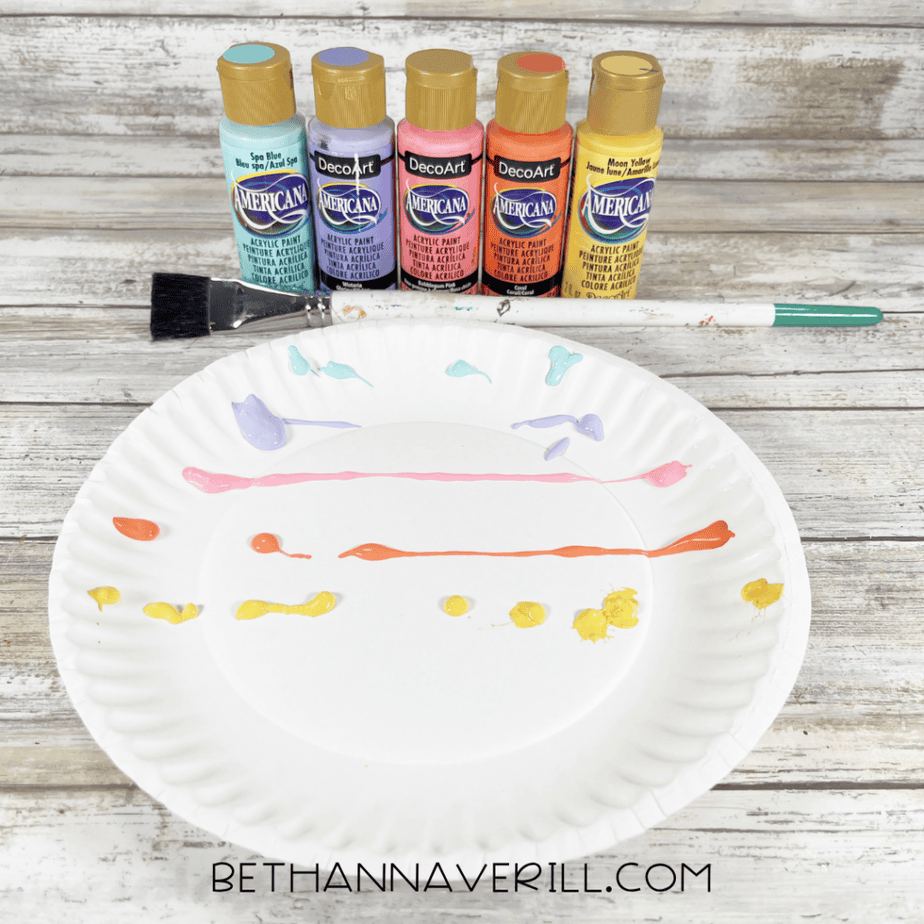 Paper plate with lines of pastel paint applied, paint bottles and brush behind it.