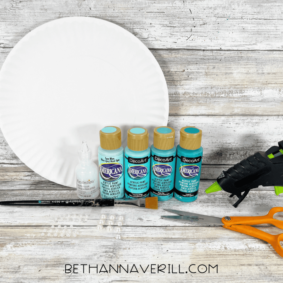 Craft supplies for a mermaid tail paper plate craft including a paper plate, acrylic paint bottles, paintbrush, glue, scissors, hot glue gun, and pearl beads.