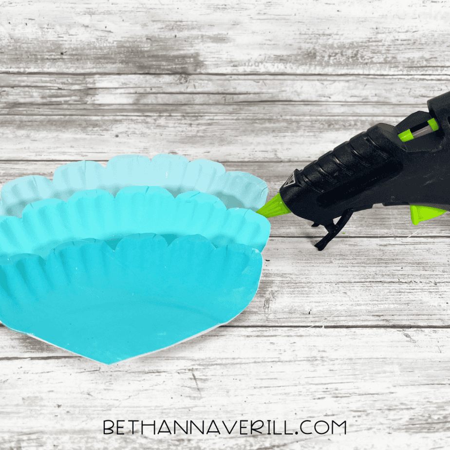 Hot glue being applied to attach turquoise painted paper plate pieces for assembling a mermaid tail craft.