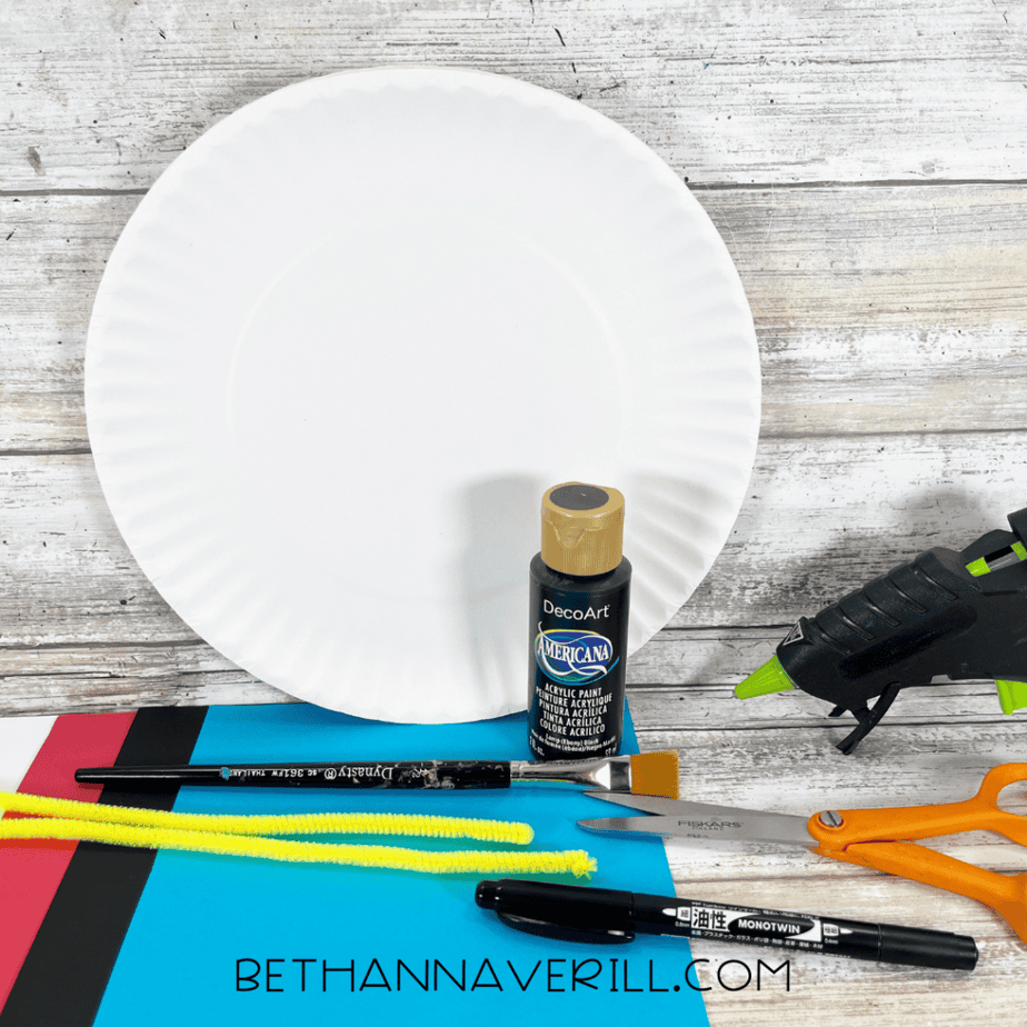 Supplies for pirate hat paper plate craft including paper plate, paint, pipe cleaner, cardstock, scissors, glue gun, and markers.