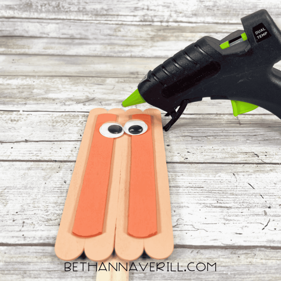 Two painted craft sticks glued vertically onto a peach-colored stick base with googly eyes being added; a hot glue gun nearby.