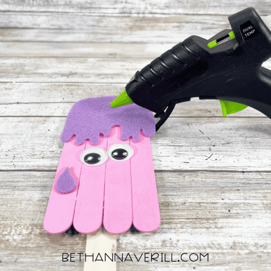 A pink painted stick popsicle base with googly eyes, a purple felt “drip” top being glued on, and a small felt piece attached to the side.