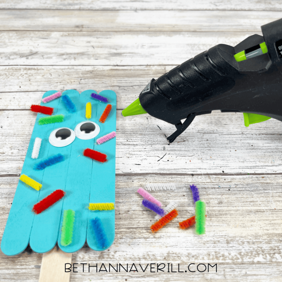 A blue popsicle stick craft covered with multicolored pipe cleaner pieces and googly eyes; glue gun nearby.