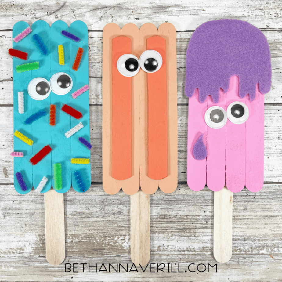 Three completed popsicle stick crafts with handles: one blue with colorful “sprinkles,” one peach with two vertical accents, and one pink with a purple felt top and side detail.