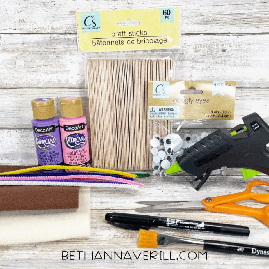 Assorted craft supplies including wooden craft sticks, googly eyes, acrylic paint bottles, pipe cleaners, felt sheets, scissors, a paintbrush, a marker, and a hot glue gun on a wooden surface.