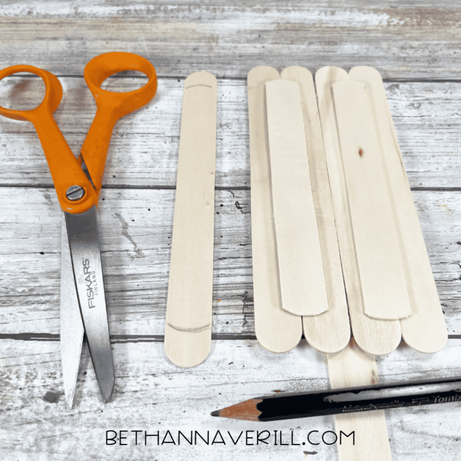 A single craft stick marked with curved cut lines at both ends next to a partially assembled stick rectangle and scissors.