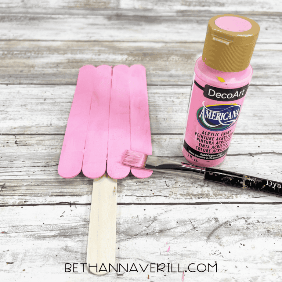 A stick rectangle attached to a vertical craft stick handle, being painted pink with a paintbrush and a bottle of pink acrylic paint.