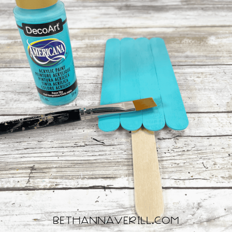 A similar stick rectangle attached to a handle, painted blue with a paintbrush and a bottle of blue acrylic paint.