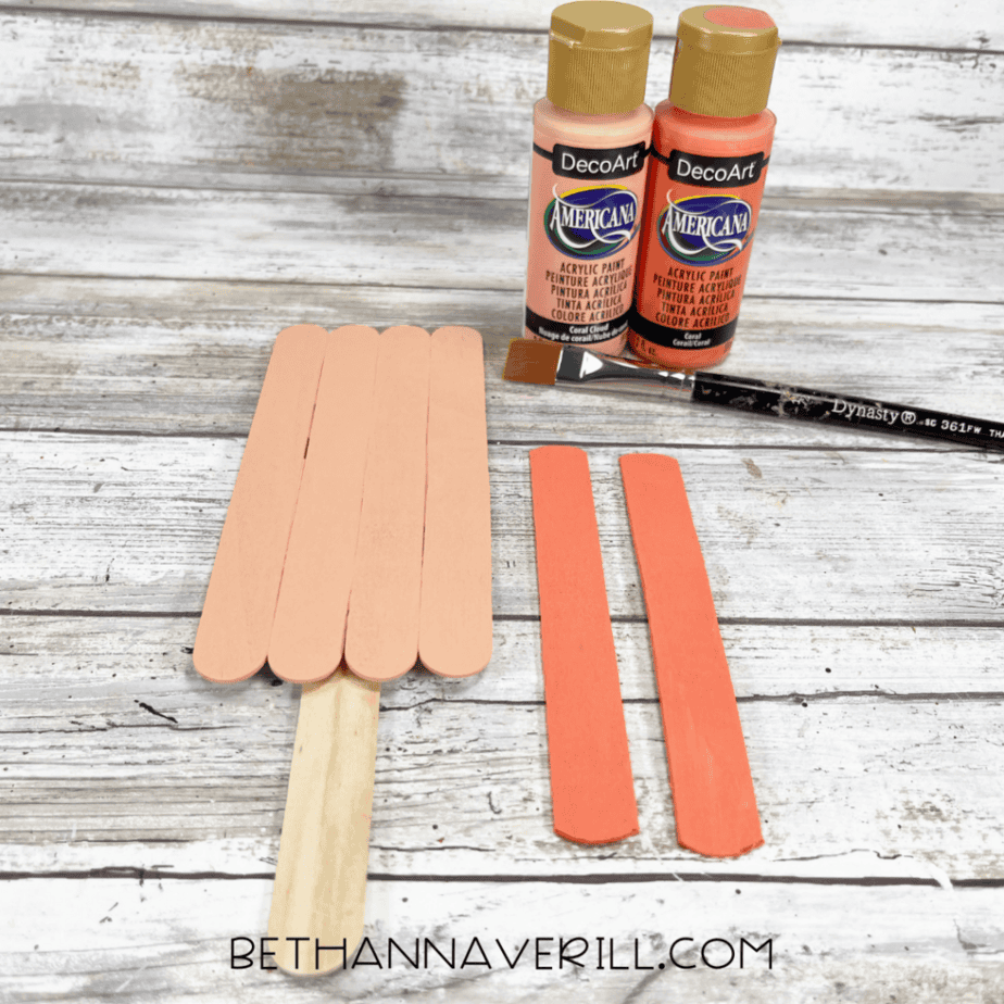 A stick rectangle attached to a handle painted a light peach color, with two darker peach-painted craft sticks placed beside it and paint bottles in the background.
