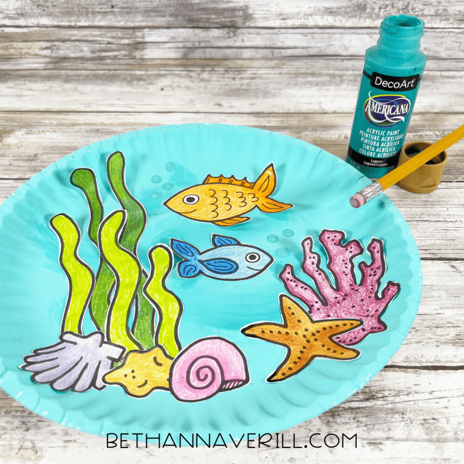 Blue painted paper plate with colored ocean-themed cutouts, small painted bubbles, an open paint bottle, and a pencil nearby.