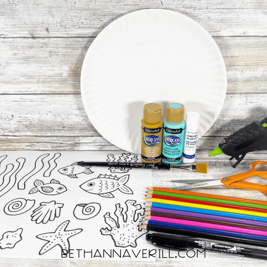White paper plate, paint bottles, glue stick, hot glue gun, scissors, paintbrush, and printed ocean-themed coloring page on a wooden surface.