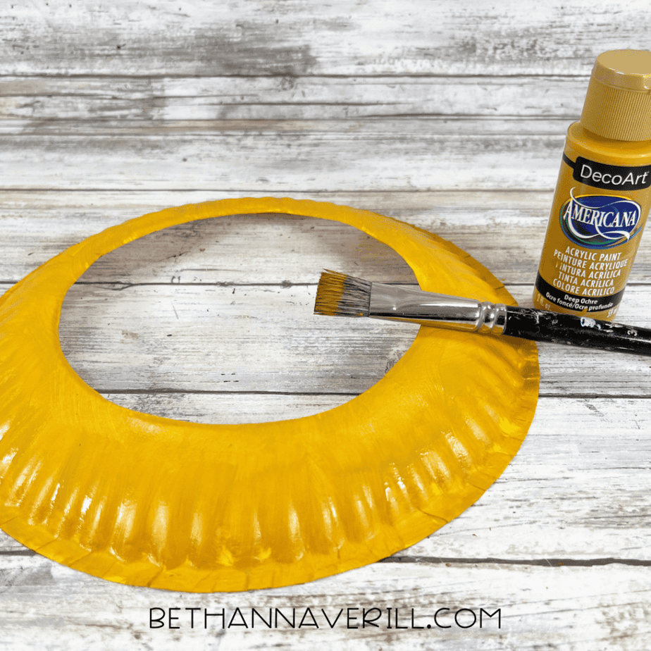 Paper plate ring painted yellow with a paintbrush resting across it and a paint bottle nearby.