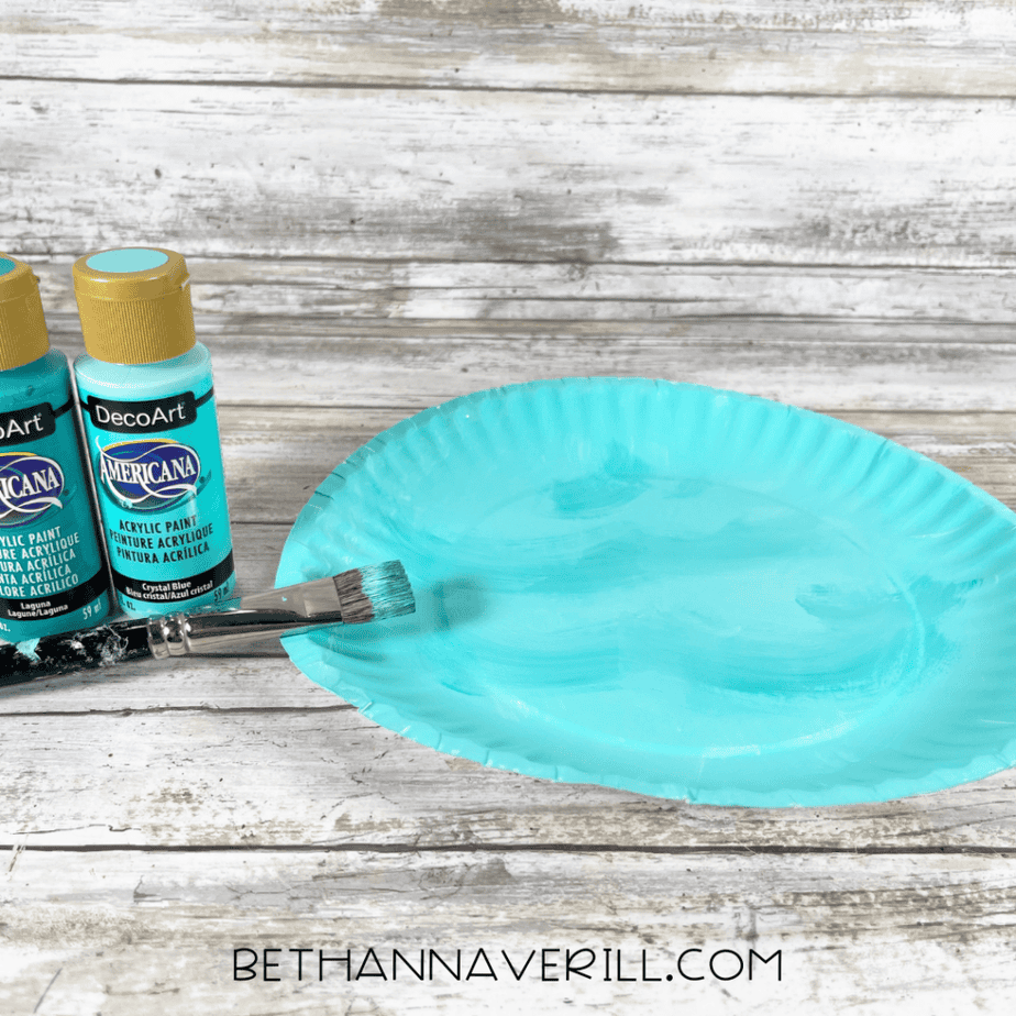 Paper plate painted light blue with a paintbrush and paint bottles next to it.