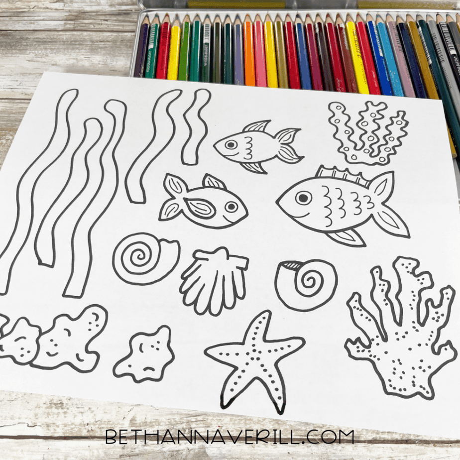 Black-and-white ocean-themed coloring sheet with fish, seaweed, shells, and coral, with colored pencils above it.