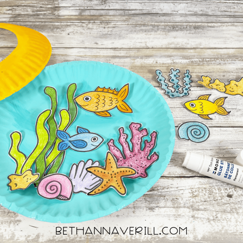 Blue-painted plate with cut-out ocean shapes being arranged inside, glue stick nearby.