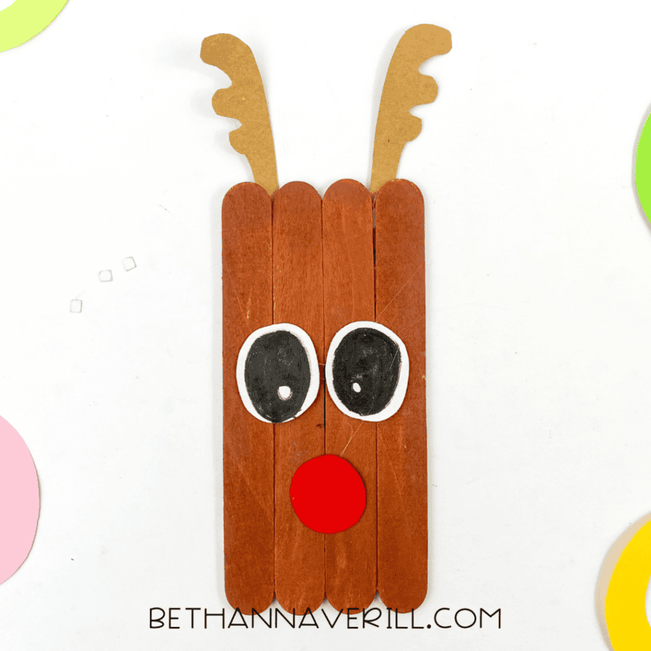 Close-up of the finished reindeer popsicle stick craft with antlers, large cartoon-style eyes, and a red nose on a white background with colorful paper edges visible.