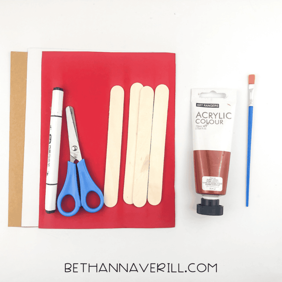Craft supplies including red, white, and brown paper, wooden craft sticks, scissors, a black marker, brown acrylic paint, and a paintbrush arranged on a white background.