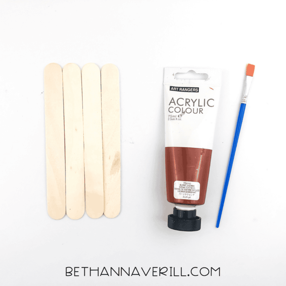Four wooden craft sticks beside brown acrylic paint and a paintbrush on a white background.