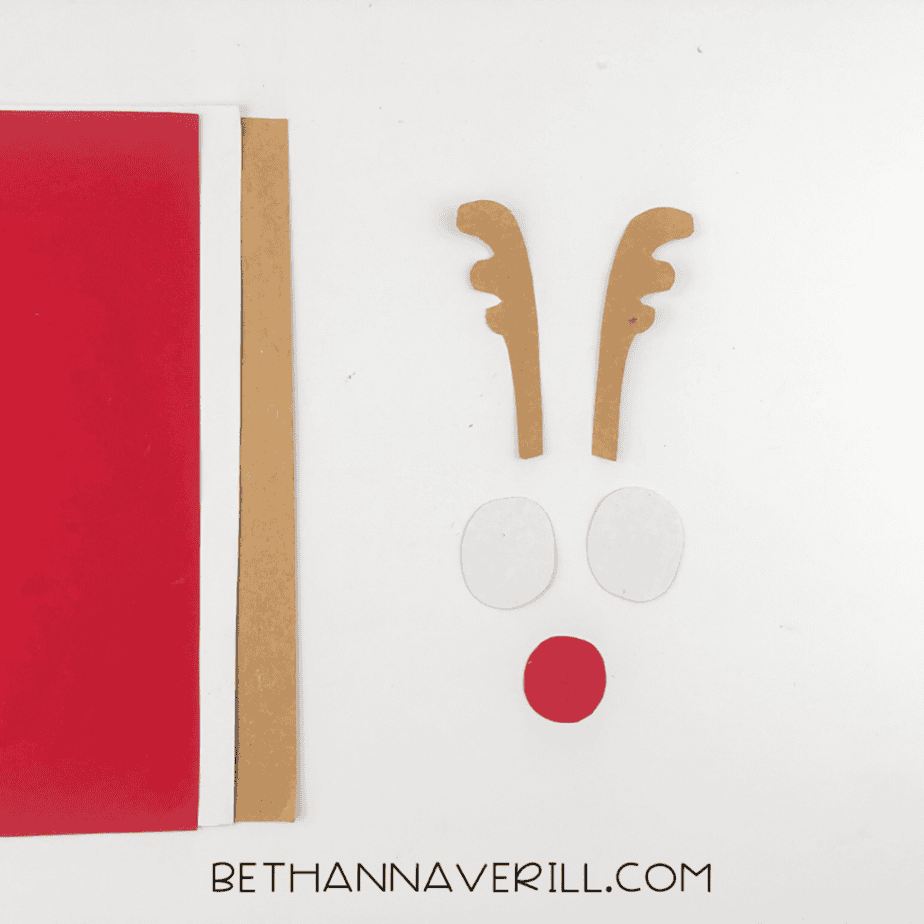Brown paper antler cutouts, white paper eye circles, and a red paper nose circle beside sheets of colored paper on a white background.